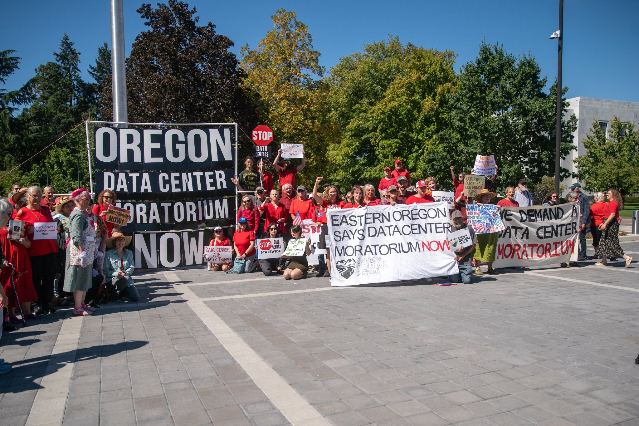 A crowd of people dressed in red stands outside a public building holding banners that read "Oregon data center moratorium now" and "Eastern Oregon says data center moratorium now.